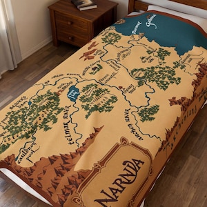 May include: A tan throw blanket featuring a map of Narnia with brown, green, and blue accents. The map includes place names like Telmar River, Witch's Camp, and Paravel. The word "Narnia" is prominently displayed.