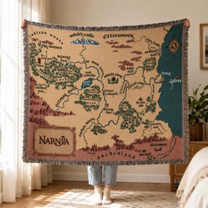 May include: A beige throw blanket featuring a map of Narnia with various locations like Witch's Castle and Cair Paravel. The map is detailed with rivers, forests, and text labels in a vintage style. The blanket has a fringed edge.