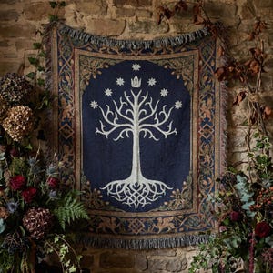 Könnte beinhalten: Eine gewebte Wandteppichdecke mit dunkelblauem Zentrum und einem weißen Baum von Gondor-Design, gekrönt von einer Krone und sieben Sternen. Der Rand zeigt komplizierte goldene und blaue Muster mit Fransen an den oberen und unteren Kanten.