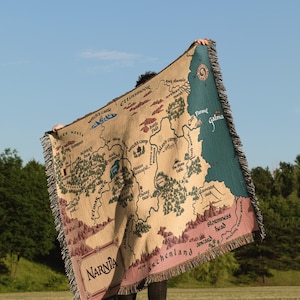 May include: A beige throw blanket featuring a map of Narnia with the text "Narnia" at the bottom. The map includes locations like Witch's Castle, Ettinsmoor, and Galma. The blanket has a fringed edge and is held up against a blue sky.