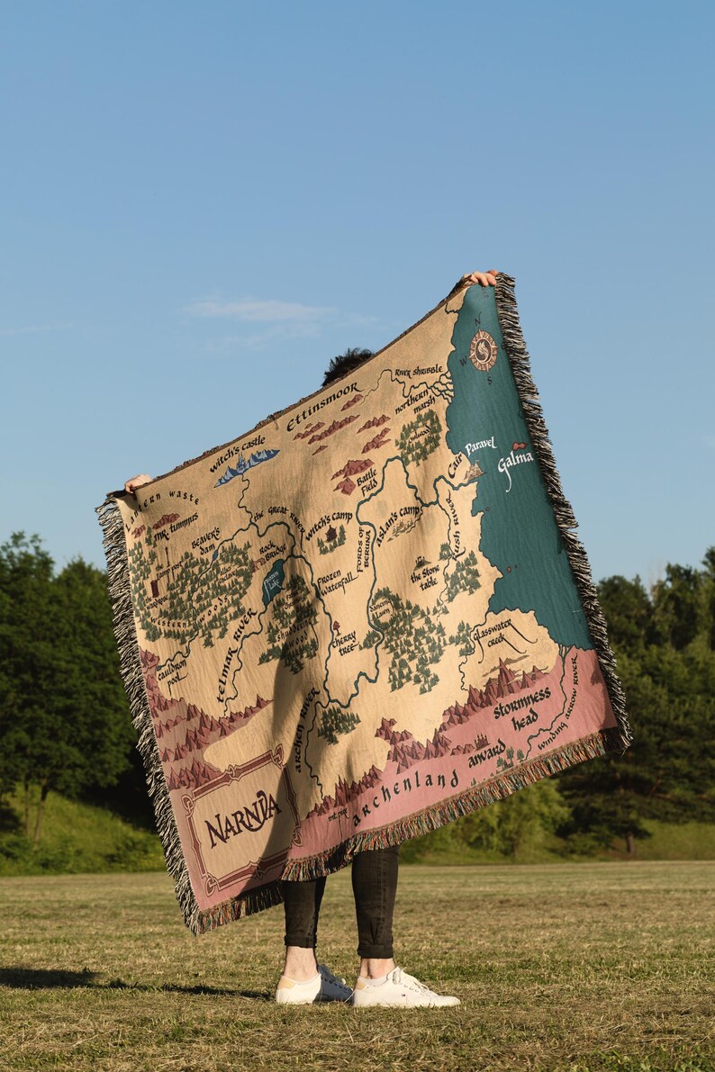 May include: A beige throw blanket featuring a map of Narnia with the text "Narnia" at the bottom. The map includes locations like Witch's Castle, Ettinsmoor, and Galma. The blanket has a fringed edge and is held up against a blue sky.