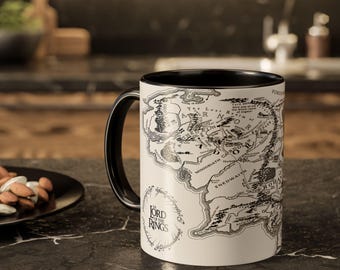 May include: A white ceramic mug with a black handle and interior. The mug features a detailed map design in black and white, with the words "Lord of the Rings" printed on the side. The mug is on a dark surface.