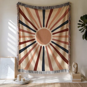 May include: A woven throw blanket with a sunburst design. The sun is a light brown circle, with rays in shades of brown, rust, and navy blue. The blanket has a fringed edge and is hanging on a wall.