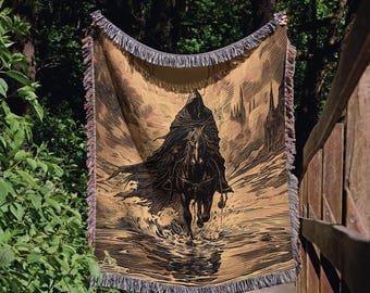 Op de afbeelding: Een geweven plaid met een zwart-bruine illustratie van een figuur met een kap op een paard in het water. Het ontwerp bevat hoge, puntige structuren op de achtergrond en een decoratieve franje langs de boven- en onderrand.