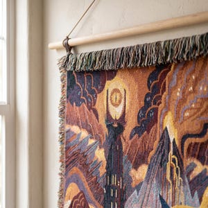 May include: A woven wall tapestry featuring a dark tower topped with a glowing yellow eye, set against a landscape of jagged mountains and clouds in shades of orange, purple, and brown. The textile hangs from a light-colored wooden rod.
