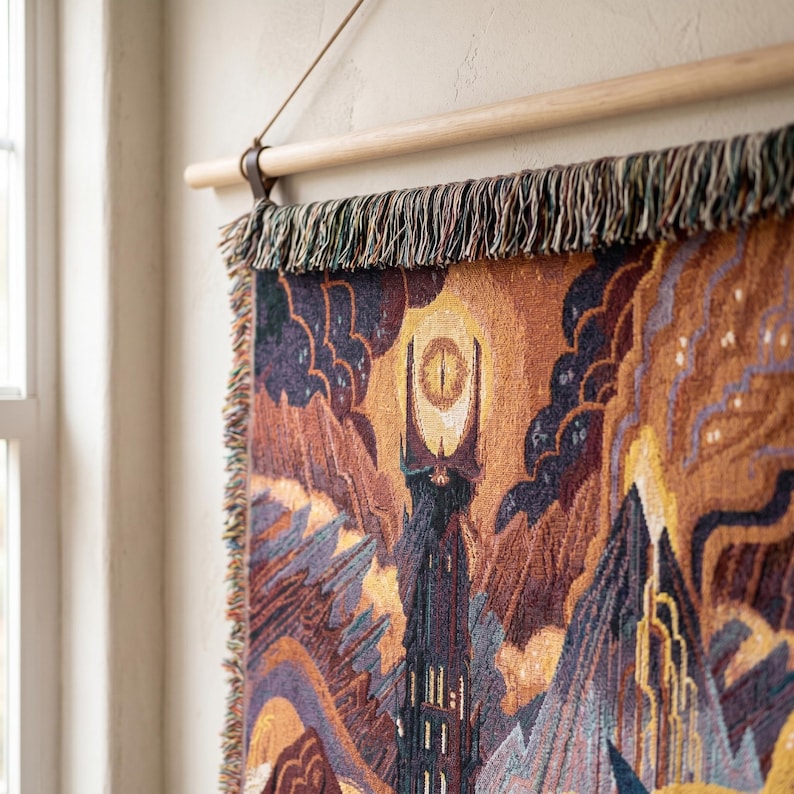 May include: A woven wall tapestry featuring a dark tower topped with a glowing yellow eye, set against a landscape of jagged mountains and clouds in shades of orange, purple, and brown. The textile hangs from a light-colored wooden rod.