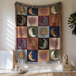 May include: A woven throw blanket featuring a repeating pattern of suns and crescent moons in a grid. The celestial design includes various colors like orange, blue, and purple. The blanket has a fringed edge and is displayed on a wall.