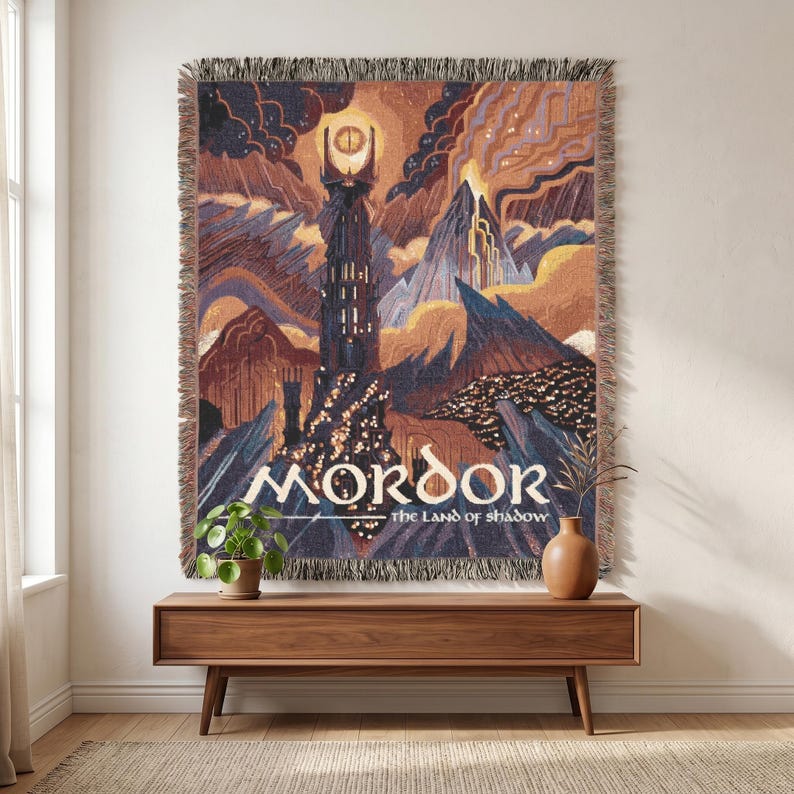 May include: A woven tapestry blanket featuring a dark fantasy landscape with a tall tower topped by a glowing eye. The scene includes jagged mountains, volcanic clouds, and the text MORDOR THE LAND OF SHADOW. The blanket has a fringed edge.