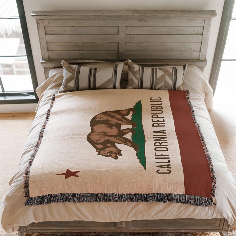 May include: A woven throw blanket featuring the California Republic flag design, including a brown bear, a green patch of grass, a red star, and the text CALIFORNIA REPUBLIC, finished with a fringed edge on a bed with two striped pillows.