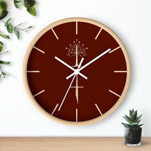 May include: A round wall clock with a light wood frame and a deep maroon face. The clock features white hour markers and white hands. Centered on the face is a white graphic illustration of a stylized tree above a broken sword design.