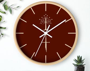 May include: A round wall clock with a light wood frame and a deep maroon face. The clock features white hour markers and white hands. Centered on the face is a white graphic illustration of a stylized tree above a broken sword design.