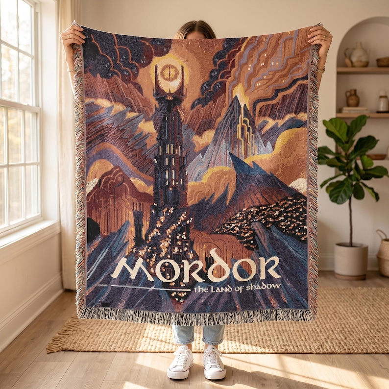 May include: A woven throw blanket featuring a dark fantasy landscape with a tall, dark tower topped by a glowing eye. The scene includes jagged mountains, swirling clouds, and the text MORDOR the Land of Shadow in a stylized white font.