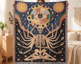 May include: A woven throw blanket featuring a dark blue background with a central skeleton figure in a seated pose with multiple arms. Above the skeleton is a large eye surrounded by moon phases, flanked by two suns, stars, and rainbow arches.