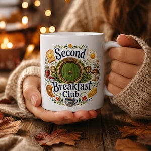 Puede incluir: Taza de cerámica blanca con la inscripción "Second Breakfast Club" y una ilustración de una puerta verde. La taza está decorada con ilustraciones de alimentos como tostadas, huevos y un cruasán. La taza es sostenida por una persona.