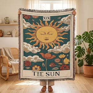 May include: A woven throw blanket featuring a tarot card design of The Sun. The center displays a large yellow sun with a face, surrounded by white clouds, red and yellow flowers, and a teal background. The text XIX and THE SUN are printed.