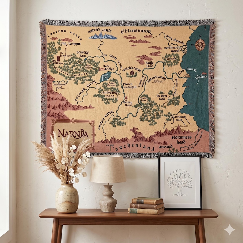 May include: A tapestry depicting a map of Narnia, with detailed illustrations of locations like Witch's Castle and Cair Paravel. The tapestry is beige, with brown and green accents. A wooden table holds a vase with dried flowers, a lamp, books, and a framed tree drawing.