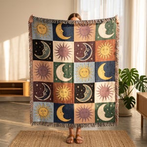 May include: A woven throw blanket featuring a repeating pattern of suns, moons, and stars in a patchwork design. The squares are in various colors including blue, green, and brown. The blanket has a fringed edge.