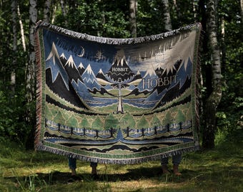 May include: A woven throw blanket featuring a landscape illustration inspired by The Hobbit, with blue mountains, green forests, and a river. The text The Hobbit and J.R.R. Tolkien is woven into the design, which has a fringed edge and blue, green, and white colors.