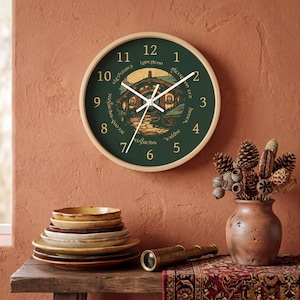 May include: A round wall clock with a light wood frame and a dark green face. The clock features gold numbers 1 through 12, white hands, and a central illustration of a round-door house. Text around the center includes Breakfast, Second Breakfast, Elevenses, Luncheon, Afternoon Tea, Dinner, and Supper.
