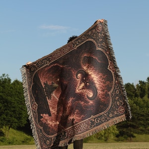 Op de afbeelding: Een rechthoekige geweven plaid met een donkere, vurige illustratie van een gehoornd wezen met gloeiende ogen te midden van wervelende rode en oranje wolken, omlijst door een decoratieve rand met bloemmotieven en franjes aan alle zijden.
