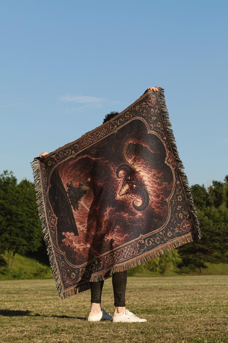 Op de afbeelding: Een rechthoekige geweven plaid met een donkere, vurige illustratie van een gehoornd wezen met gloeiende ogen te midden van wervelende rode en oranje wolken, omlijst door een decoratieve rand met bloemmotieven en franjes aan alle zijden.