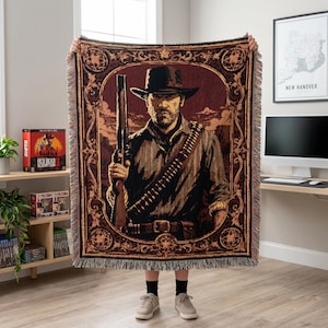 Può includere: Una coperta tessuta che raffigura il ritratto di un uomo con cappello da cowboy, che impugna un fucile e indossa una bandoliera. Il disegno è racchiuso in una cornice rettangolare decorata dai toni scuri con frange sui bordi.