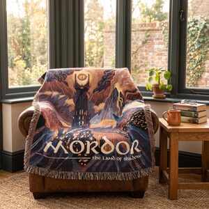May include: A woven throw blanket featuring a dark, detailed illustration of a tall tower with a glowing eye at the top, set against a mountainous landscape. The text MORDOR THE LAND OF SHADOW is printed in a stylized font across the bottom section.