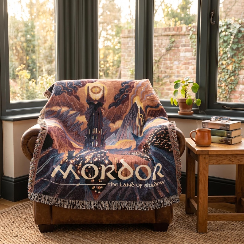 May include: A woven throw blanket featuring a dark, detailed illustration of a tall tower with a glowing eye at the top, set against a mountainous landscape. The text MORDOR THE LAND OF SHADOW is printed in a stylized font across the bottom section.