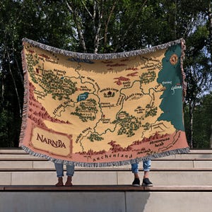 May include: A beige throw blanket featuring a map of Narnia with green, brown, and pink accents. The blanket has a fringed edge and the word "Narnia" is printed in a decorative font. The map includes place names like "Telmar River" and "Archenland".