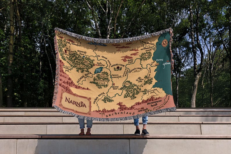 May include: A beige throw blanket featuring a map of Narnia with green, brown, and pink accents. The blanket has a fringed edge and the word "Narnia" is printed in a decorative font. The map includes place names like "Telmar River" and "Archenland".