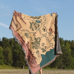 May include: A beige throw blanket with a map design, featuring green trees, blue water, and pink borders. The map includes text such as "Archen River" and "Witch's Camp". The blanket has a fringed edge and is held up against a blue sky.