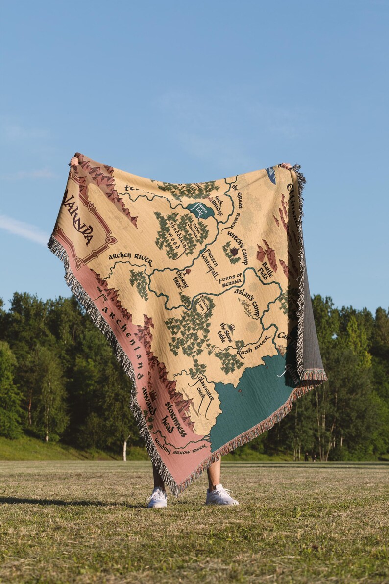 May include: A beige throw blanket with a map design, featuring green trees, blue water, and pink borders. The map includes text such as "Archen River" and "Witch's Camp". The blanket has a fringed edge and is held up against a blue sky.