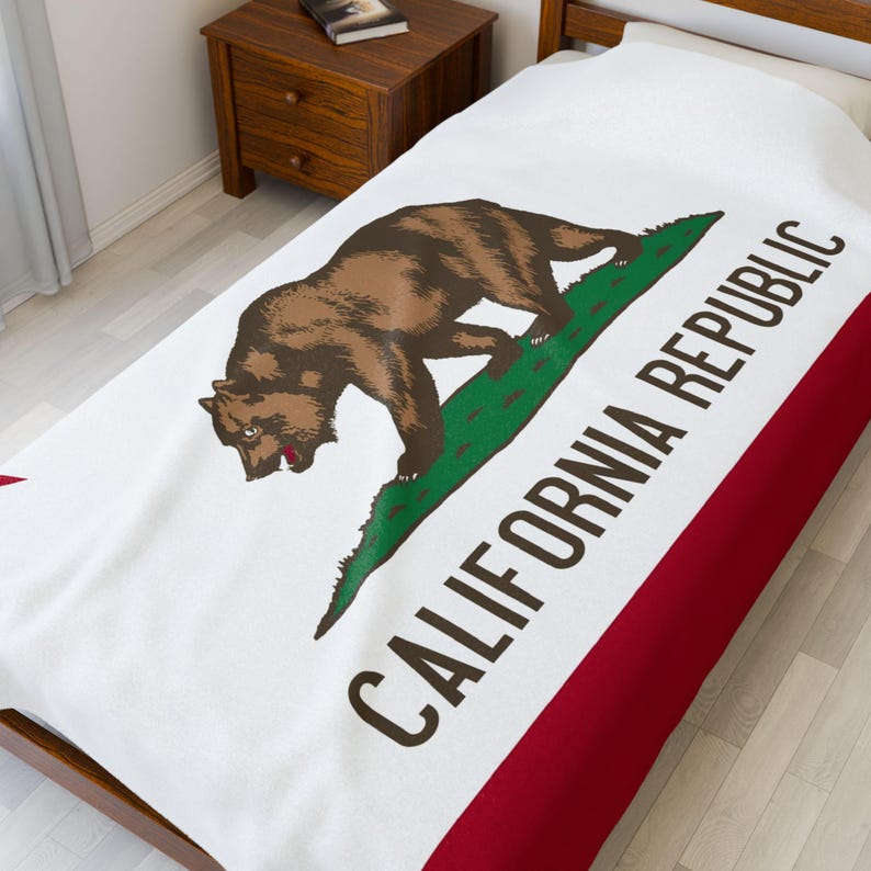 May include: A white bed blanket featuring the California state flag design, including a brown grizzly bear walking on a green patch of grass, a red star in the upper left corner, and the text CALIFORNIA REPUBLIC printed in dark brown letters.