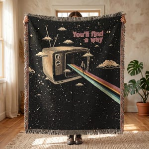 Może przedstawiać: Czarny tkany koc z grafiką telewizora retro, tęczową ścieżką i napisem "You'll find a way". Koc ma frędzelkową krawędź i wzór gwieździstego nieba.