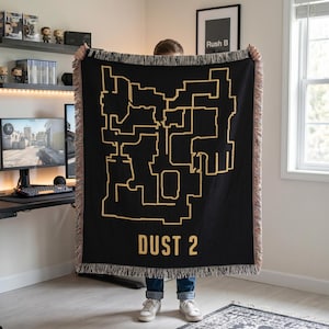 Puede incluir: Una manta tejida negra que presenta el contorno dorado del diseño del mapa Dust 2 de un videojuego. El texto DUST 2 está impreso en dorado en la parte inferior. La manta tiene un borde con flecos y un patrón de hilos multicolores.