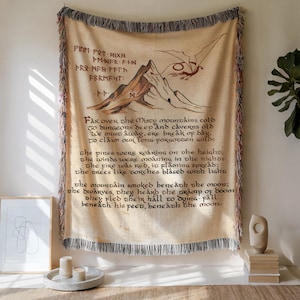 May include: Beige woven throw blanket with a mountain and dragon design. The blanket features text from a poem. The edges are fringed. The artwork is in shades of brown and red.