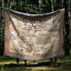 May include: A beige tapestry blanket with a detailed map design, featuring mountains, rivers, and text. The blanket has a fringed edge and is held up in front of a forest backdrop. The map includes illustrations of creatures and locations.