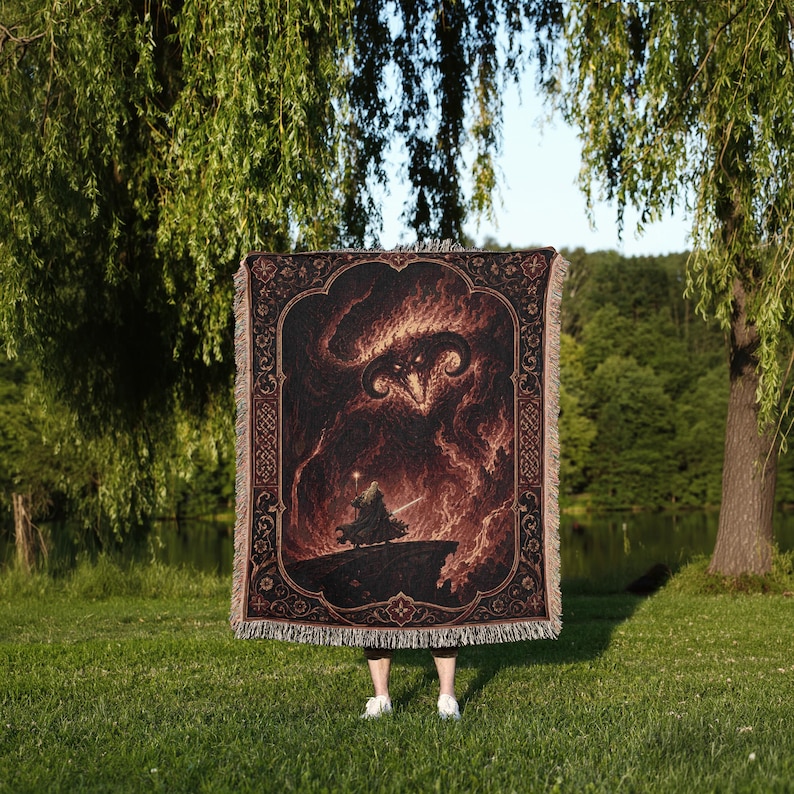 May include: A rectangular woven throw blanket featuring a dark, fiery illustration of a horned demon looming over a cloaked figure holding a staff. The design is framed by an ornate, dark border with intricate patterns and has a fringed edge.