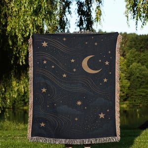May include: A dark blue woven throw blanket featuring a celestial design. The blanket has a crescent moon, stars, and flowing lines in a gold color. The edges are fringed.