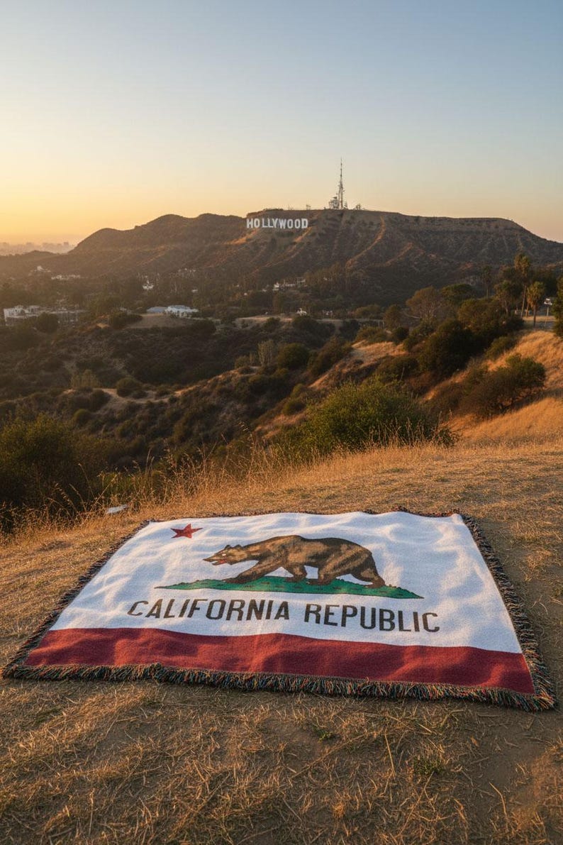 May include: A blanket with the California state flag design, featuring a bear, star, and the words "CALIFORNIA REPUBLIC." The blanket is white with a red border and is displayed outdoors with the Hollywood sign in the background.