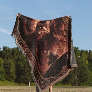 Op de afbeelding: Een geweven plaid met een donkere, vurige sc&egrave;ne met een figuur die een gloeiende staf en zwaard vasthoudt, staand op een gebogen oppervlak onder een groot, schimmig wezen. De deken heeft een decoratieve rand met ingewikkelde bloemmotieven.
