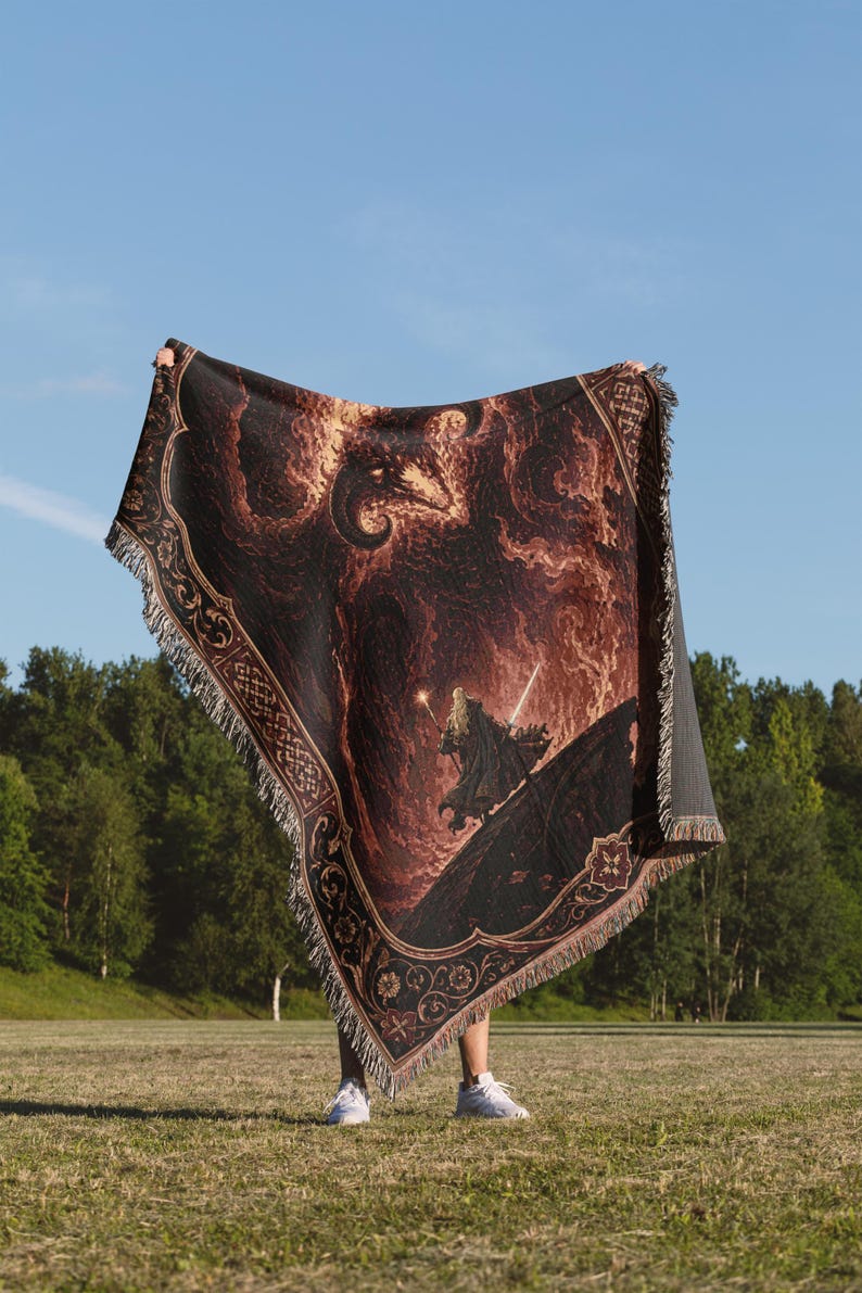 Op de afbeelding: Een geweven plaid met een donkere, vurige sc&egrave;ne met een figuur die een gloeiende staf en zwaard vasthoudt, staand op een gebogen oppervlak onder een groot, schimmig wezen. De deken heeft een decoratieve rand met ingewikkelde bloemmotieven.