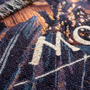May include: A woven tapestry blanket featuring a dark purple and orange color palette with a textured surface. The fabric displays a partial graphic design with the letter M and a circular shape in cream-colored thread, finished with a fringed edge.