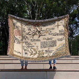 May include: A beige tapestry blanket with a map design, featuring a dragon, mountains, and text. The blanket has a fringed edge and is held up against a backdrop of trees. The map appears to be a fantasy theme.