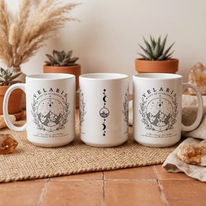 Puede incluir: Tres tazas de cerámica blanca con ilustraciones negras. Dos tazas presentan un diseño de montaña y estrella con el texto "VELARIS CITY OF STARLIGHT". La tercera taza tiene un diseño de media luna. Las tazas están sobre una estera tejida.