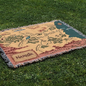 May include: A rectangular woven throw blanket featuring a map of Narnia. The blanket has a tan background with a detailed map design in shades of brown, green, and blue. The word "Narnia" is displayed in a decorative font.