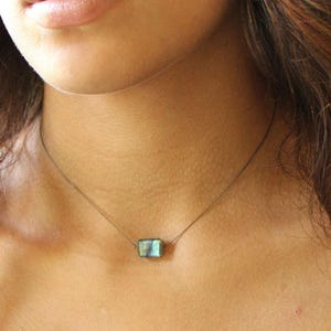 May include: A woman wearing a black top with a delicate silver chain necklace featuring a small, rectangular, iridescent green gemstone pendant.