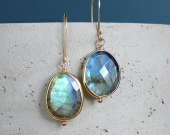 Peut inclure: Une paire de boucles d'oreilles pendantes dorées avec des pierres précieuses ovales bleu-vert iridescentes. Les boucles d'oreilles sont serties dans des lunettes dorées et ont une petite perle dorée en bas. Fond : cylindre en béton gris clair.
