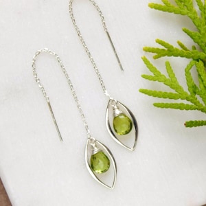 May include: A pair of silver threader earrings featuring a delicate chain that loops through the ear. Each earring has a marquise-shaped silver frame enclosing a single, faceted, pear-shaped peridot green gemstone suspended by silver wire.