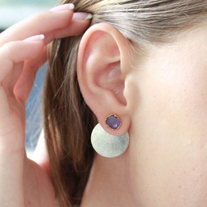 Peut inclure: Gros plan sur une oreille portant des boucles d'oreilles argentées et dorées. Chaque boucle d'oreille est composée d'une petite pierre précieuse violette ronde sertie d'or, fixée à un grand disque argenté texturé.